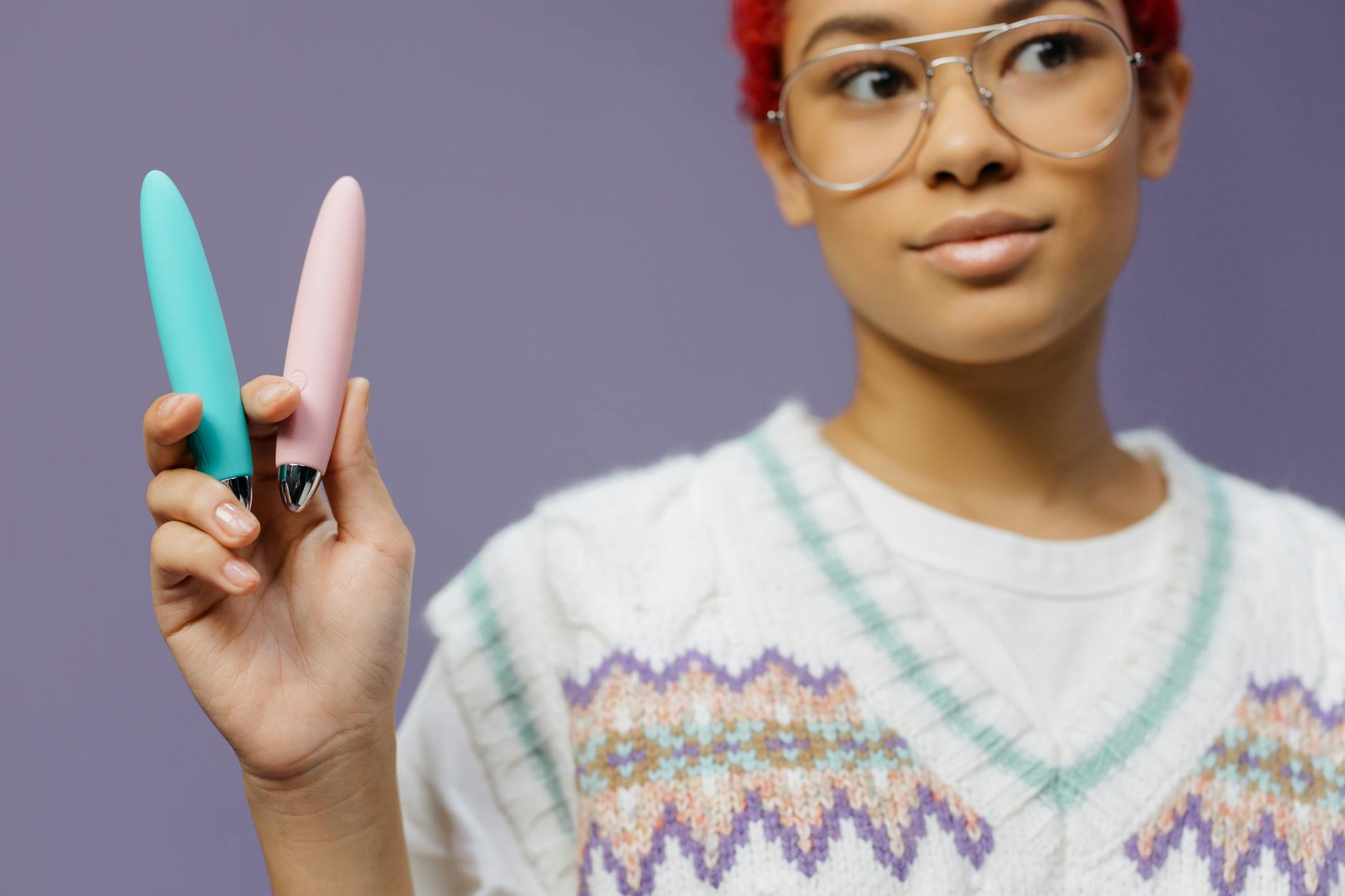 A woman holding two colorful silicone vibrators against a purple background, representing different clitoral vibrator options