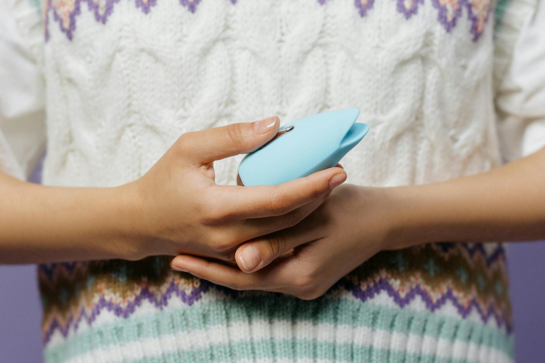 Close-up of hands holding a blue personal vibrator against a knitted sweater, representing intimate exploration.